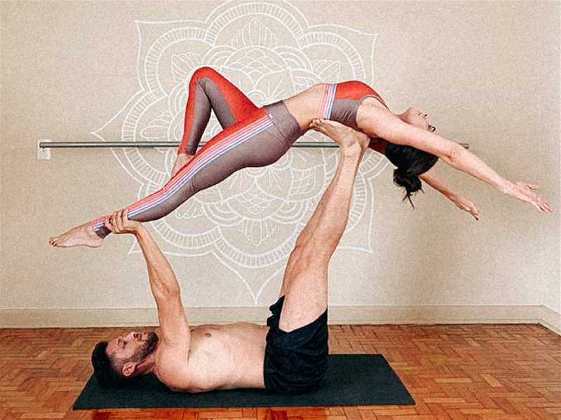 homem e mulher praticando pilates com ele deitado no chao e ela nos pés dele no alto