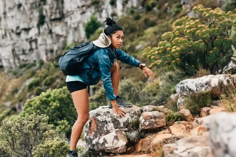 Mulher com look para trilha usando mochila, jaqueta leve e tênis, explorando terreno rochoso.